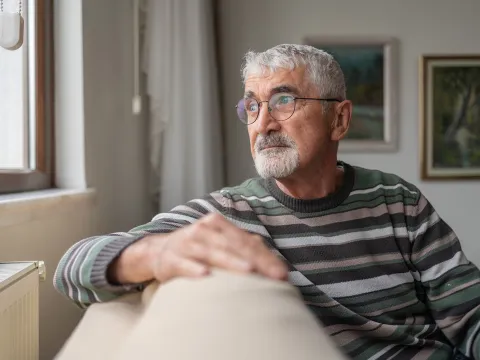 An Senior Man with gray hair and glasses is sitting on a beige couch, wearing a striped sweater.