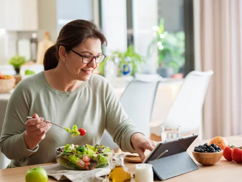 A woman reading on a tablet while eating at the table.