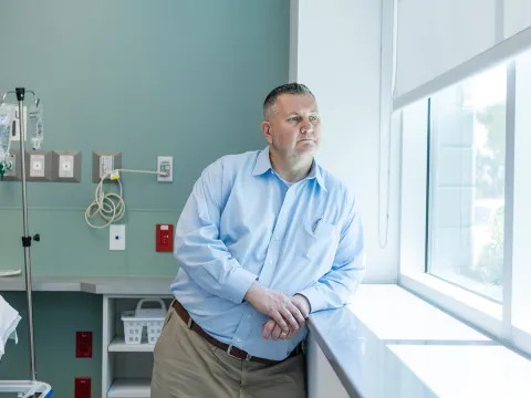 Man in blue shirt leans on counter in hospital room, gazing out window with IV stand and medical outlets in background.