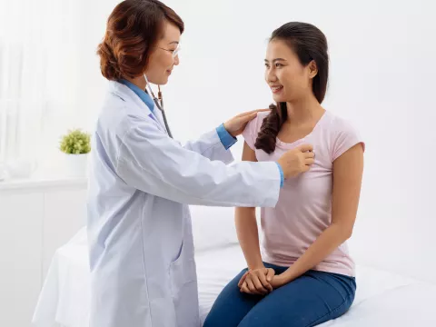 A woman is sitting on an exam table while a doctor listens to her heart.