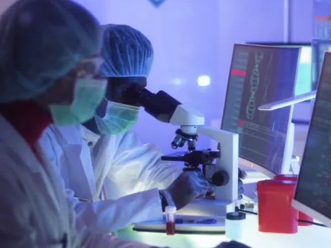 2 individuals in full ppe in a research lab face computers, one looks into a microscope