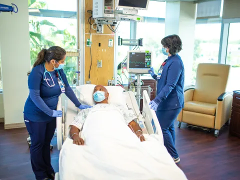 Man in the ICU talking to a nurse while another nurse checks a monitor. All are wearing masks.