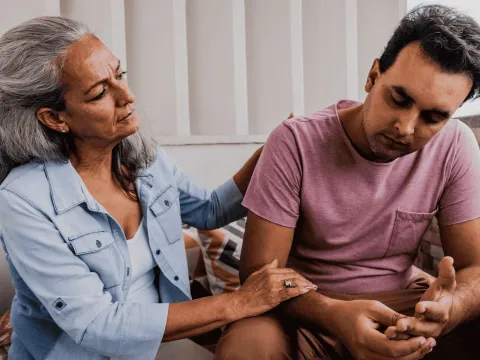 An older woman sits on the couch comforting her adult son.