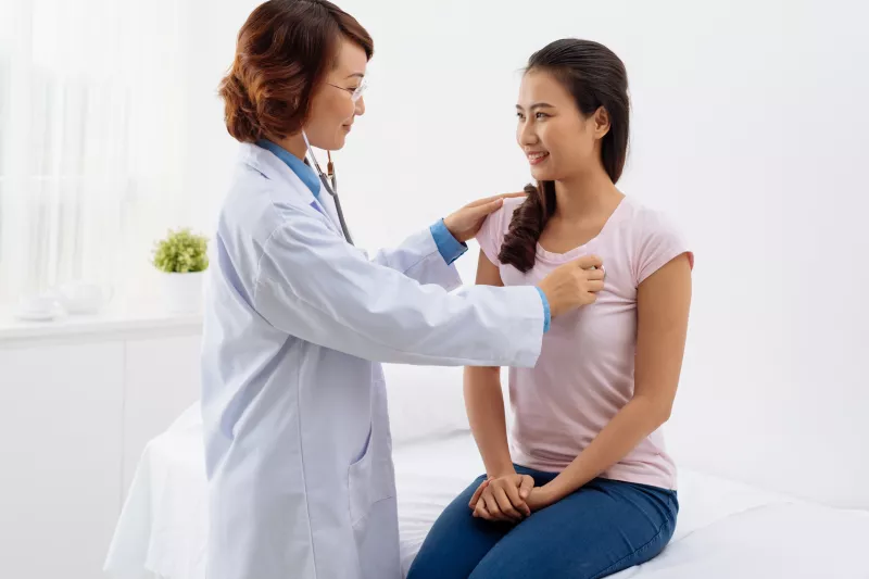 A woman is sitting on an exam table while a doctor listens to her heart.