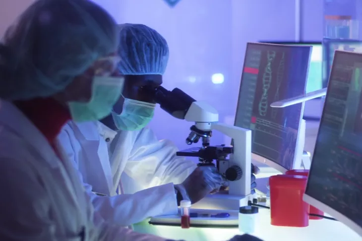 2 individuals in full ppe in a research lab face computers, one looks into a microscope