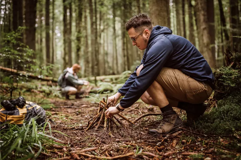 Man building a campfire in a forest while a woman collects timber in the background.