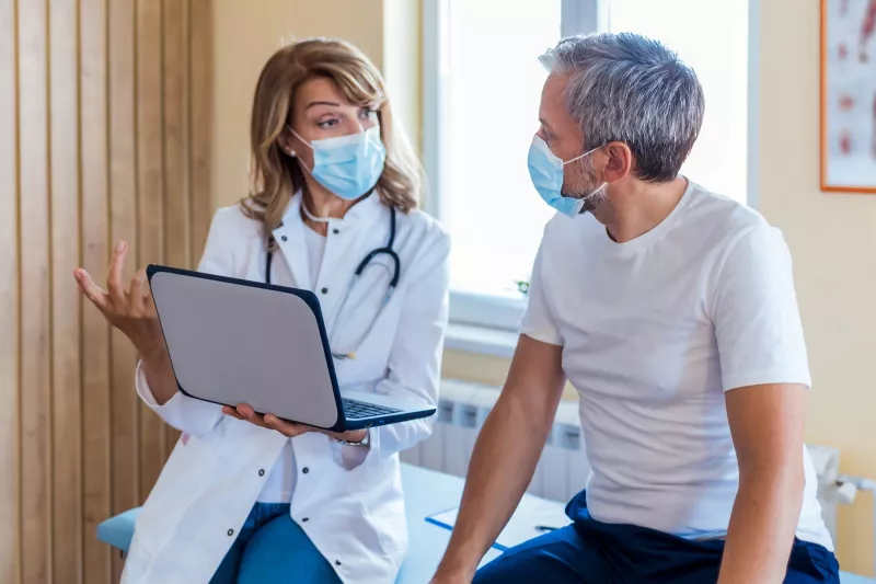 Female doctor talking with male patient holding laptop, both masked