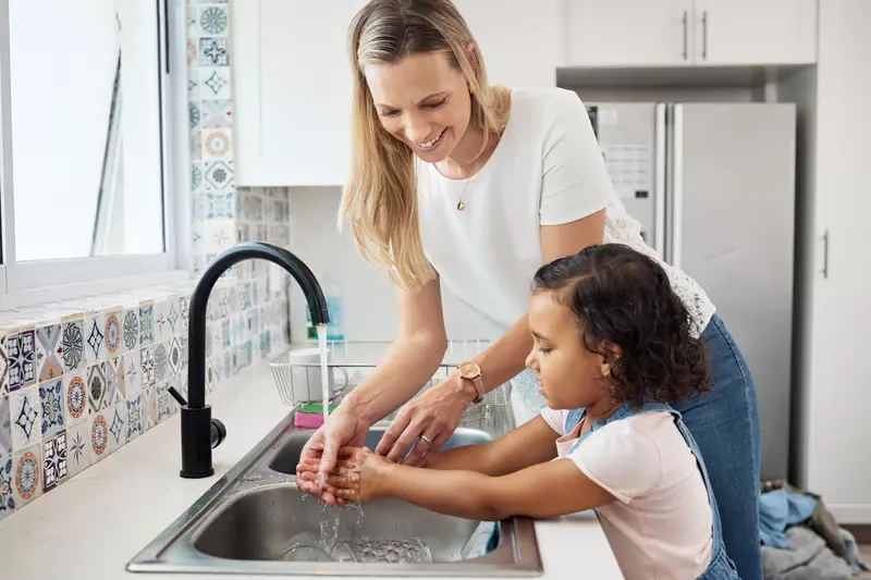 Mother showing her daughter how to wash her hands at the kitchen sink.
