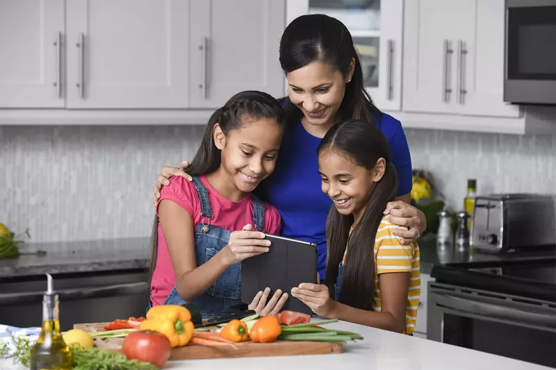 A mother and her 2 young daughters reading a recipe on a tablet in the kitchen.